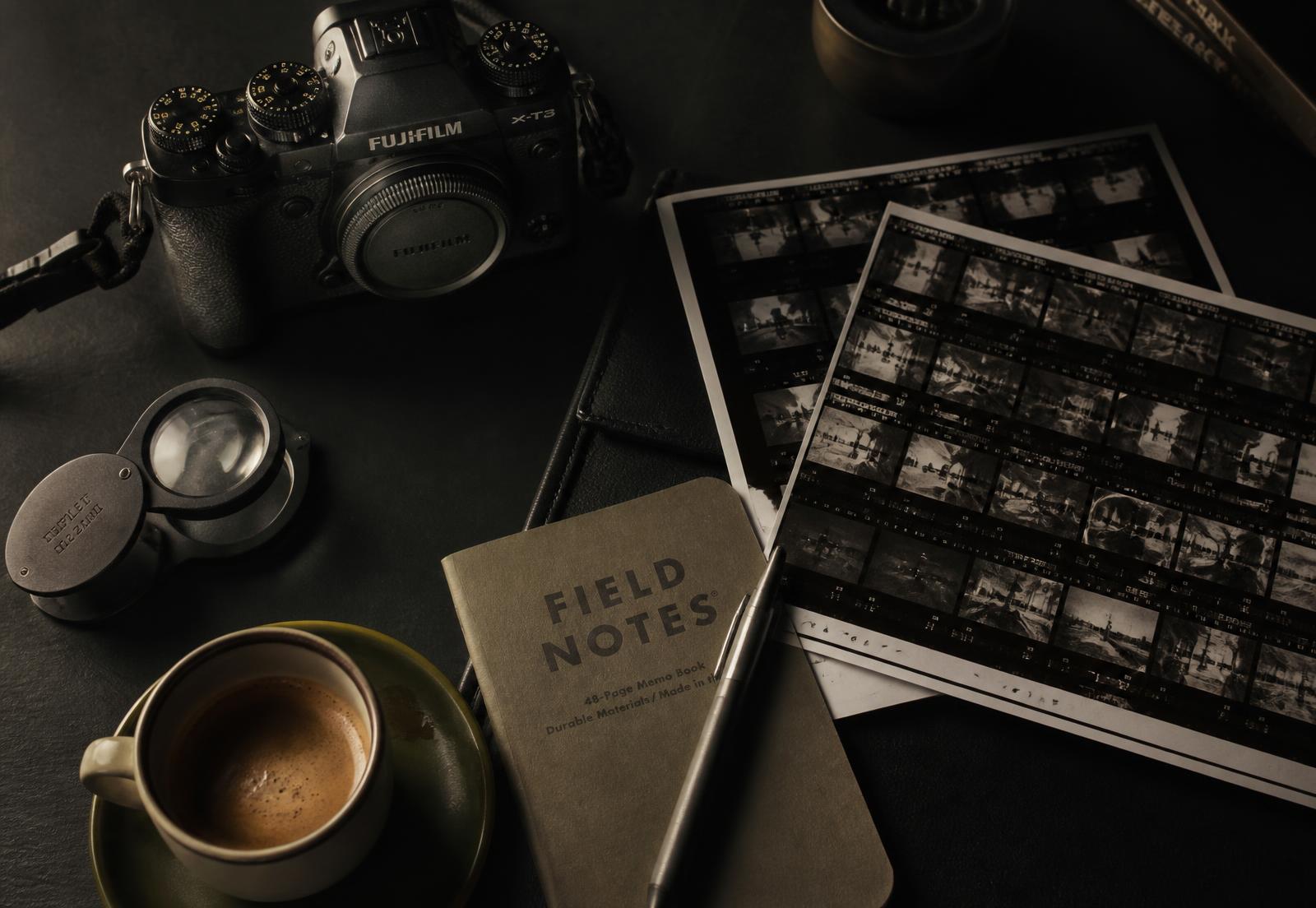 Camera, contact sheets, notebook and espresso on a dark studio desk